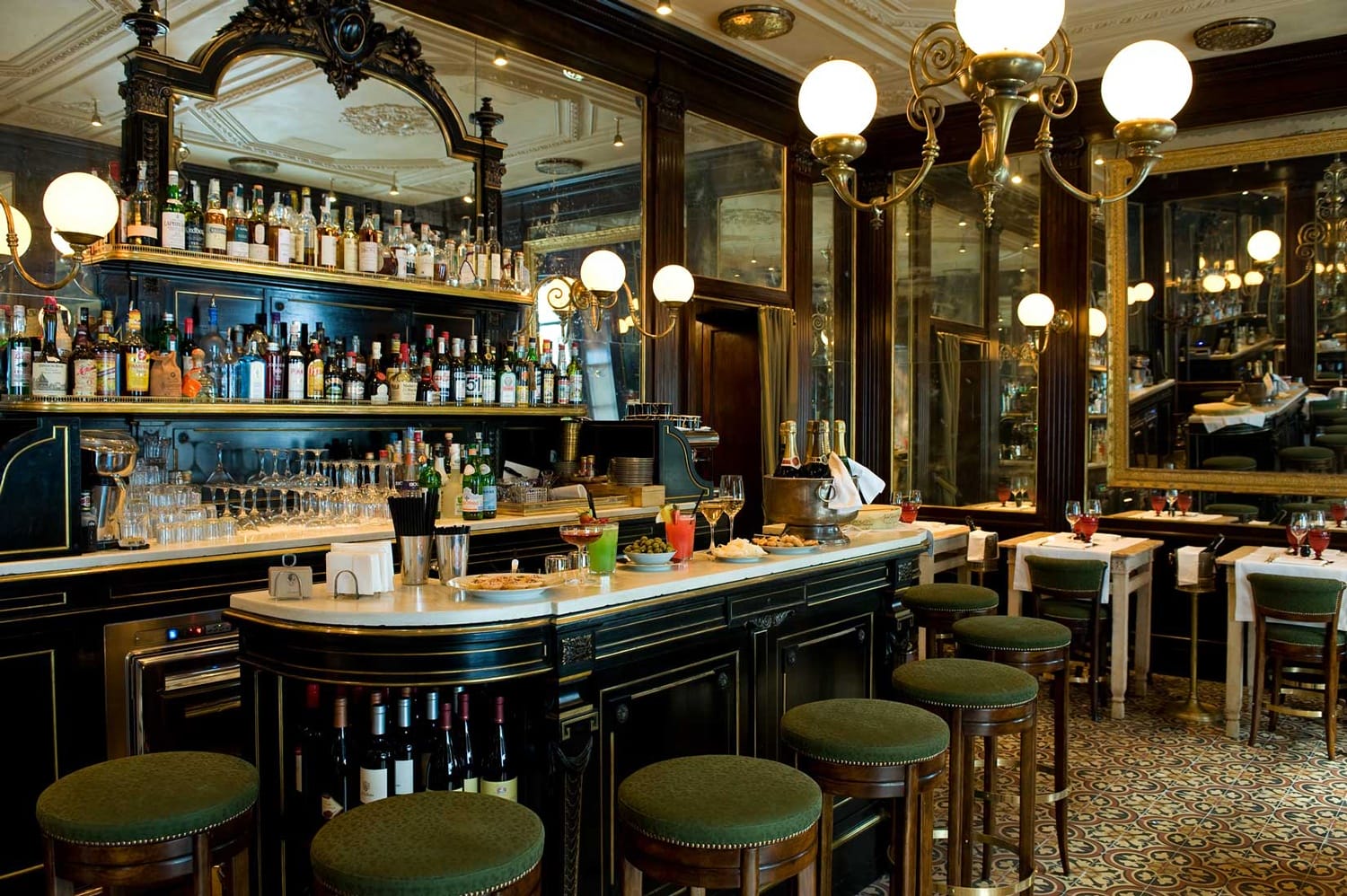 Elegant vintage bar interior with a stocked counter, ornate mirrors, and green upholstered stools.