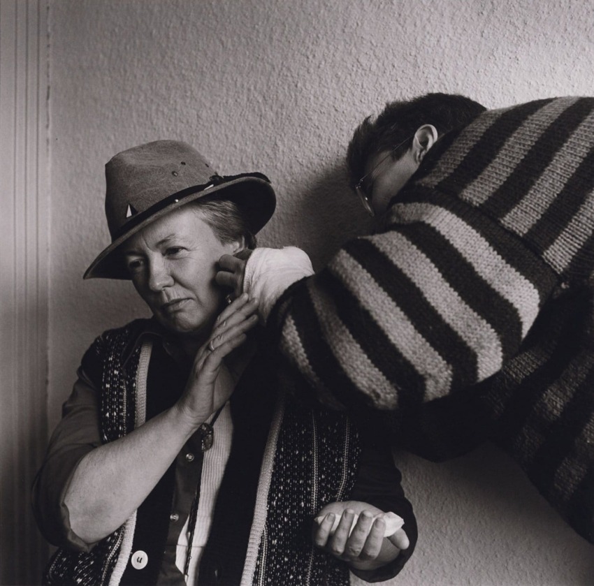 Person adjusts another person's scarf while they wear a hat, both standing indoors against a plain wall.