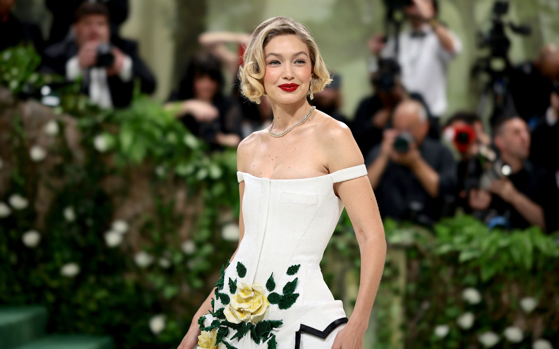 Person in an off-shoulder white dress with floral details, posing in front of photographers surrounded by greenery.
