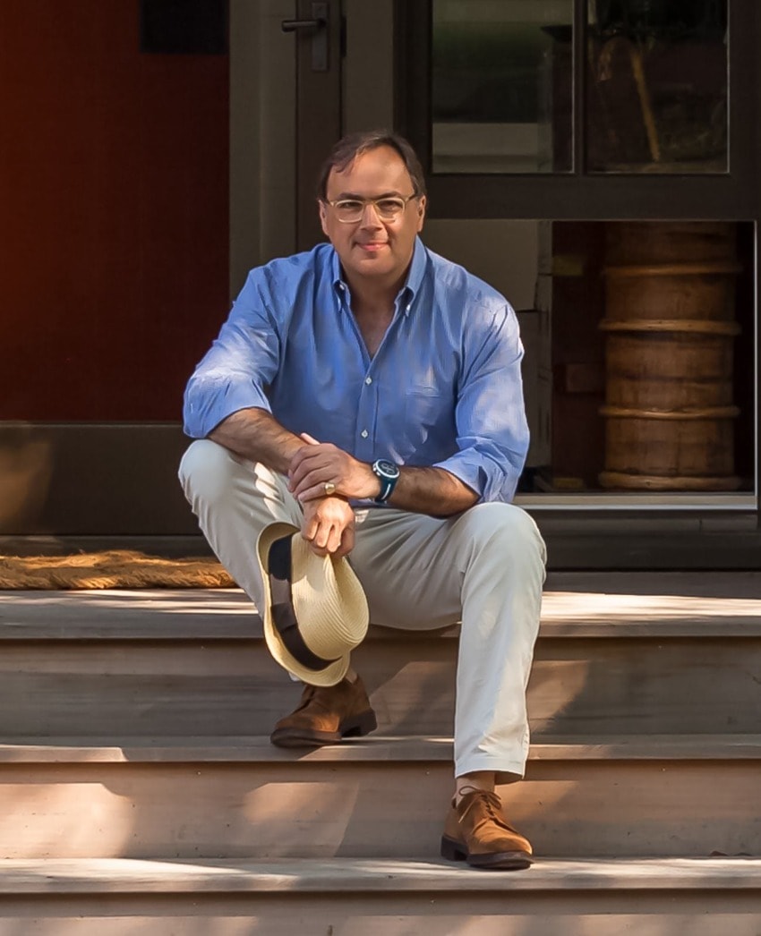 Man sitting on outdoor steps, wearing a blue shirt and holding a hat, with a relaxed pose and a slight smile.