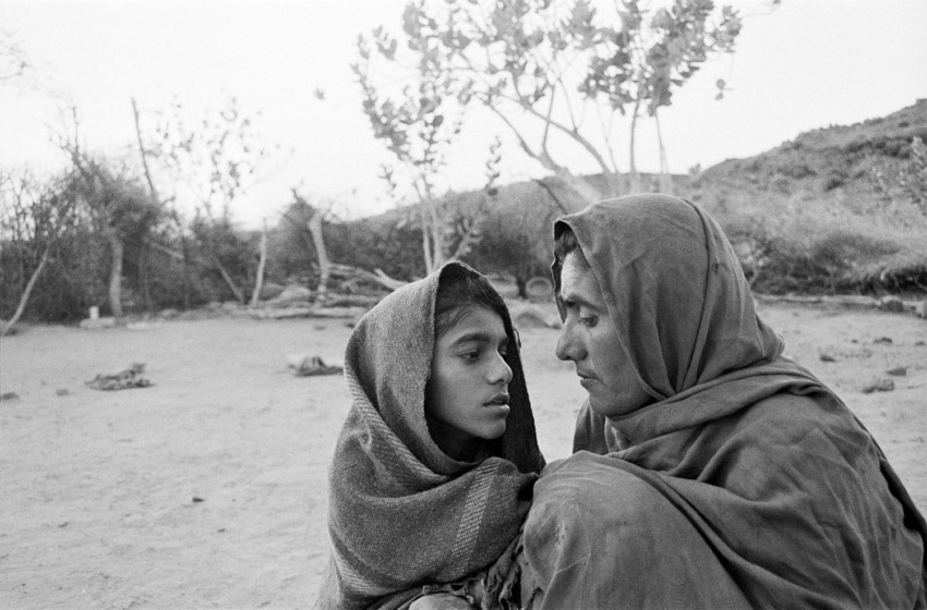 A mother and child sitting outdoors wrapped in shawls, facing each other with a serene expression in a rural setting.