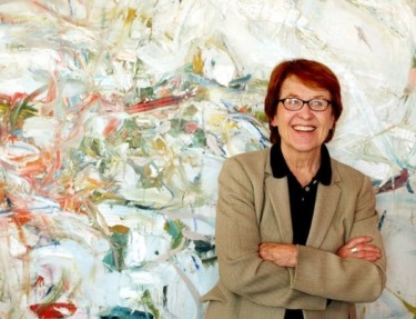 A person smiling in front of an abstract painting with colorful brushstrokes.