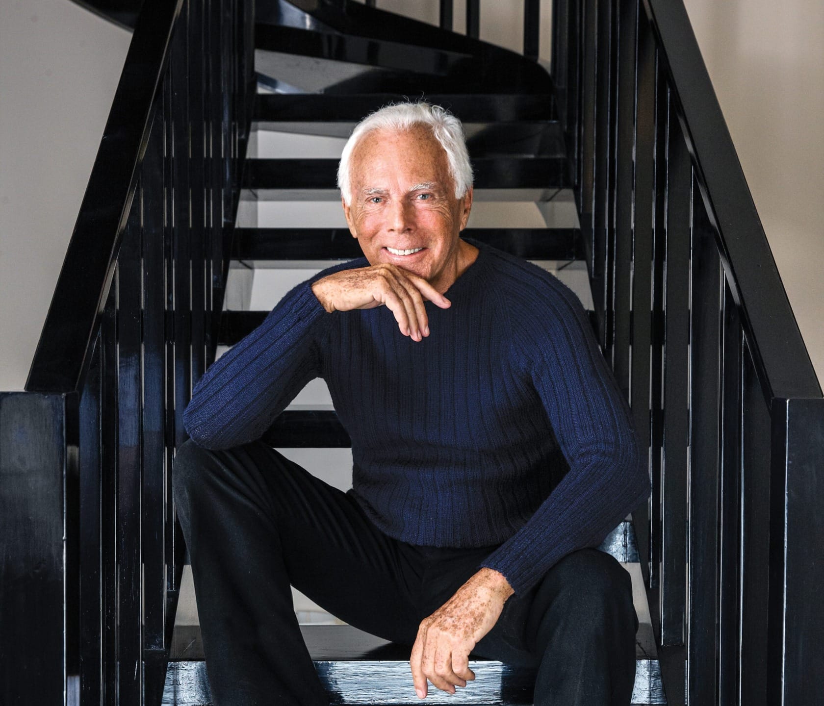 Man sitting on black staircase, wearing a dark blue sweater, smiling and resting his chin on his hand.
