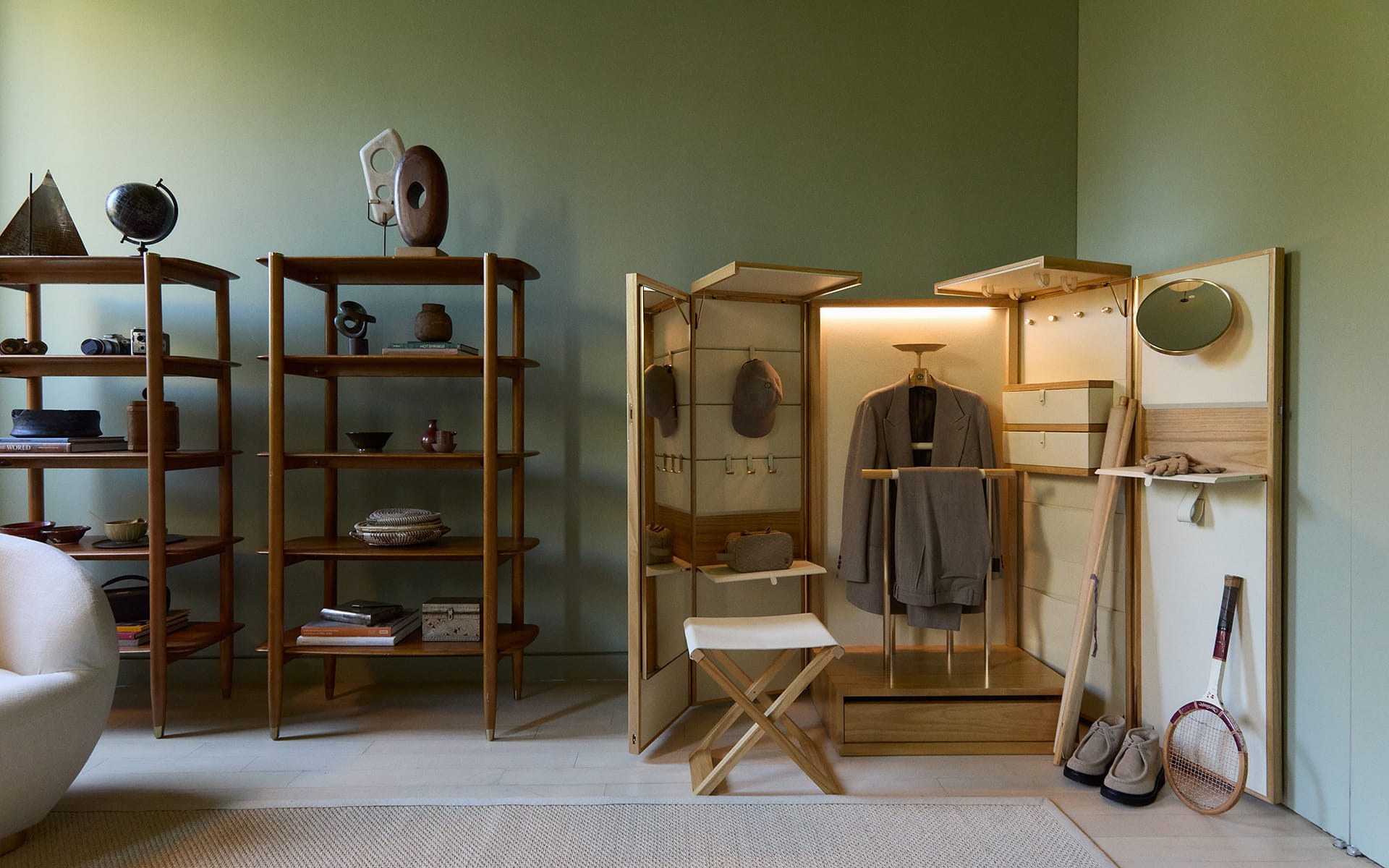 Modern room with wooden shelves displaying artifacts and a wardrobe cabinet with a hanging coat and mirror, in green tones.