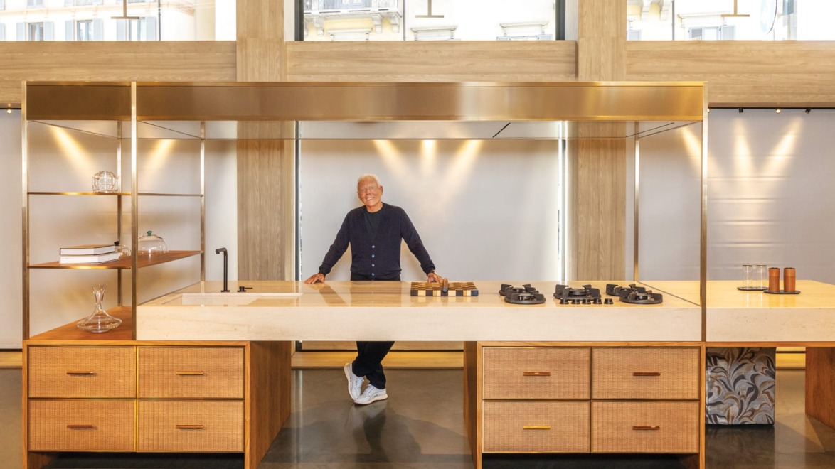 Modern kitchen with a smiling person, stylish wooden cabinets, sleek island, and gas stovetops in a bright room.