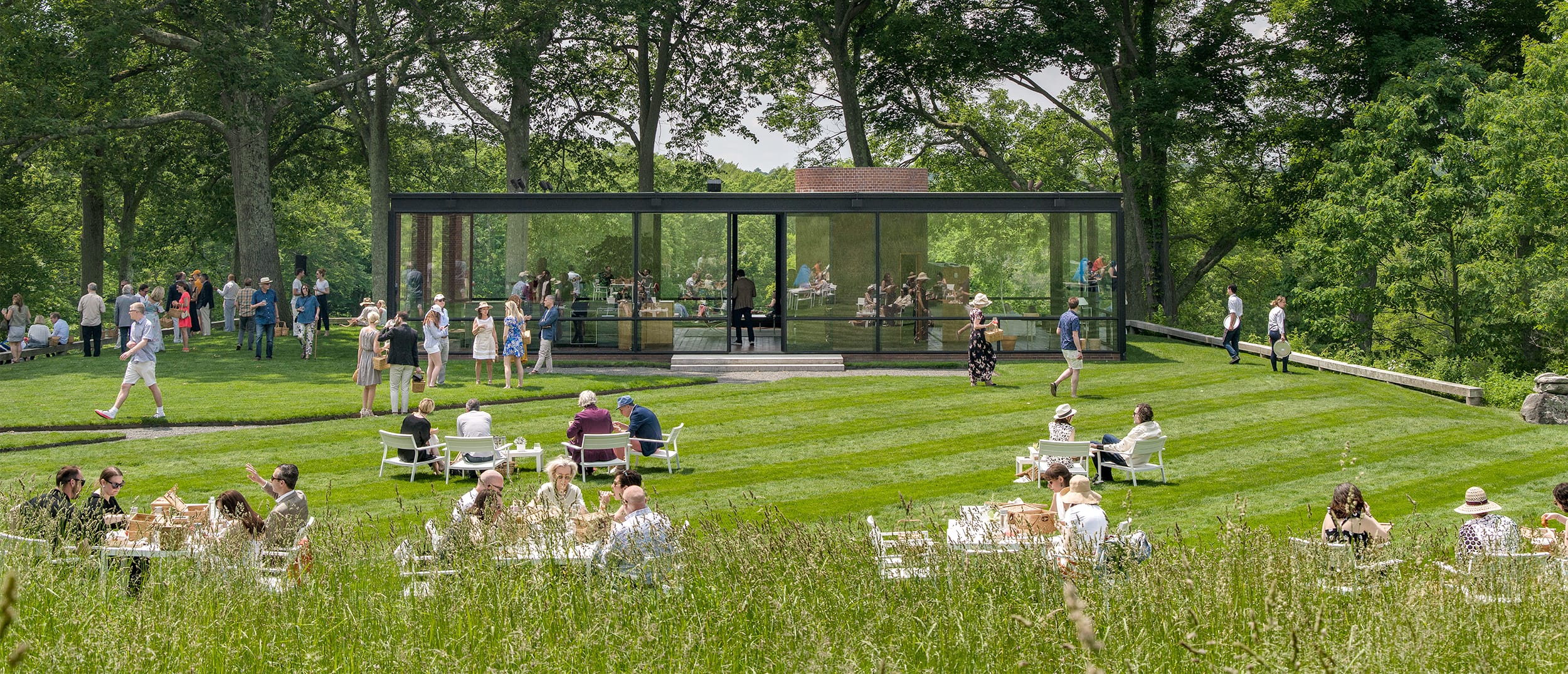 People enjoying a picnic on a grassy lawn outside a modern glass house surrounded by trees.