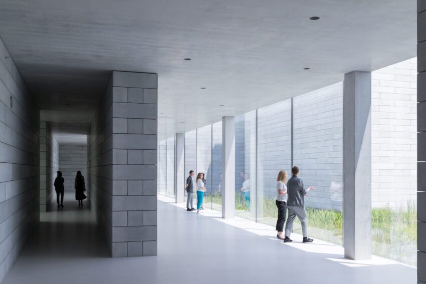 People walking and standing in a modern, minimalistic hallway with large windows displaying an outdoor view.