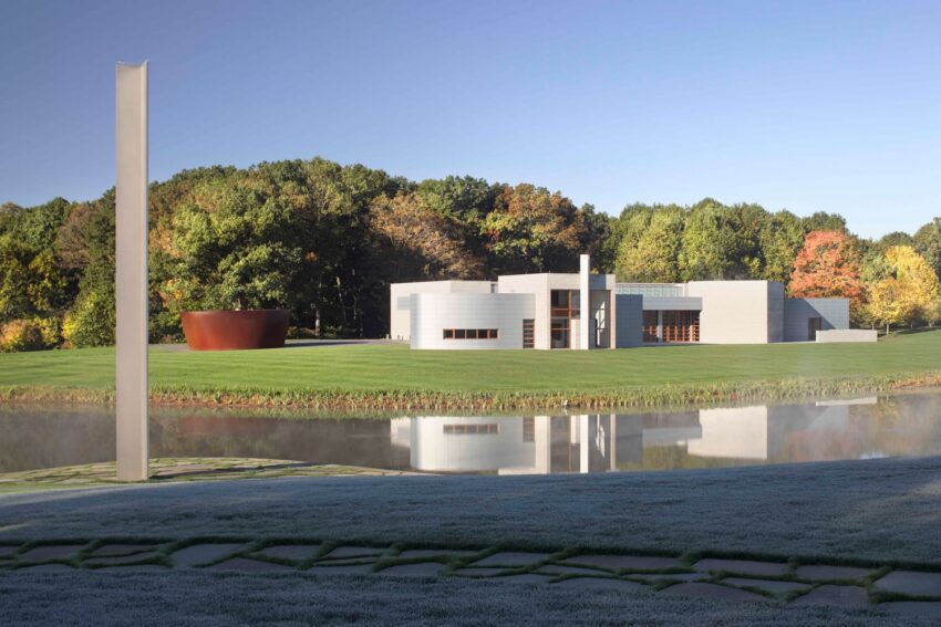 Modern white building with glass windows surrounded by trees and grass, reflecting in a pond under a clear blue sky.