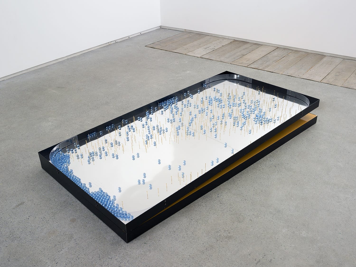 Modern art installation with a rectangular mirror base, blue balls, and vertical rods, placed on a concrete floor.