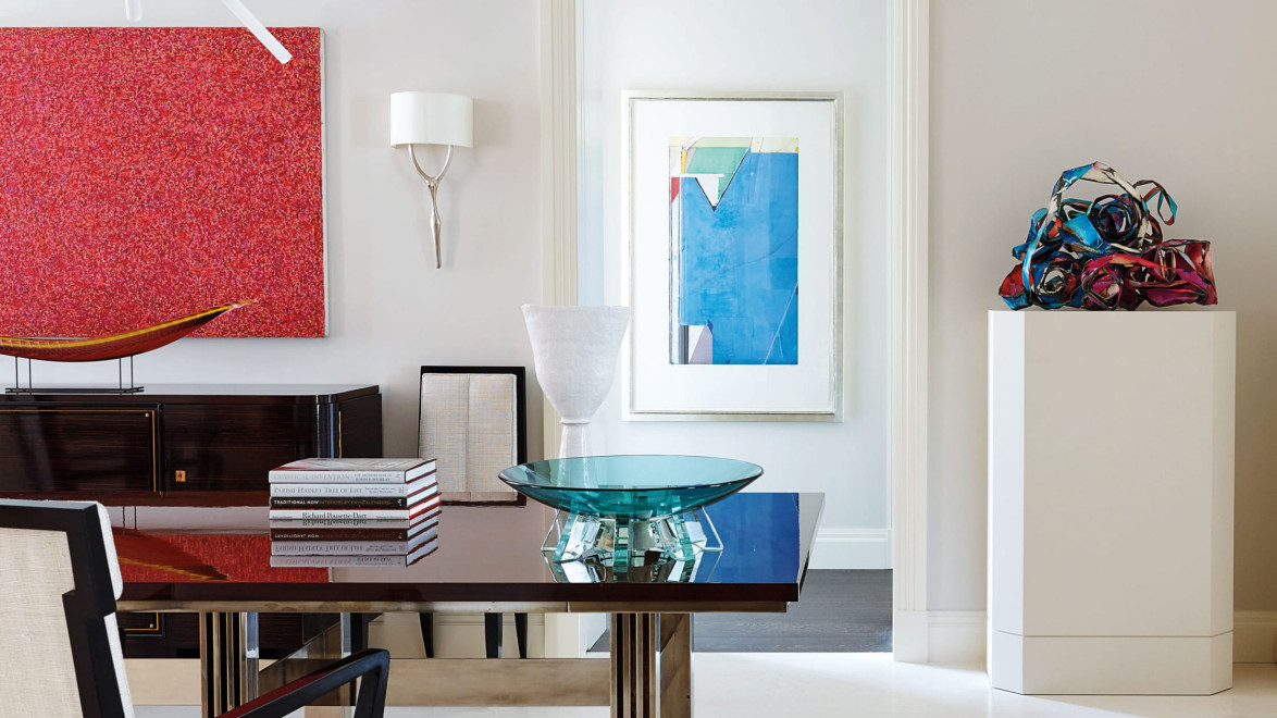 Modern art gallery interior with a sleek table, red and blue abstract paintings, stacked books, and colorful sculpture.