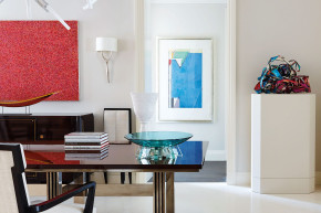 Modern art gallery interior with a sleek table, red and blue abstract paintings, stacked books, and colorful sculpture.