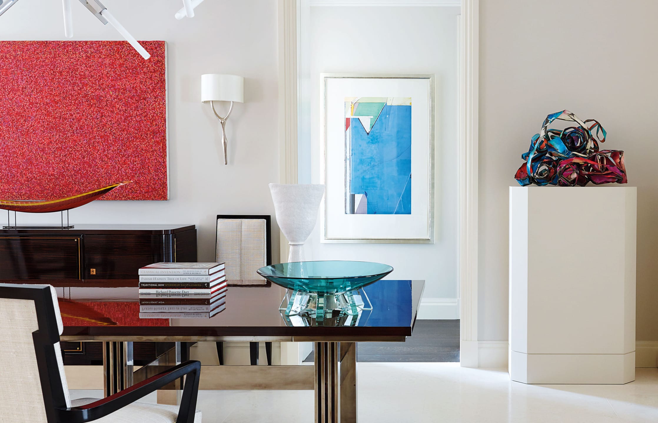 Modern art gallery interior with a sleek table, red and blue abstract paintings, stacked books, and colorful sculpture.