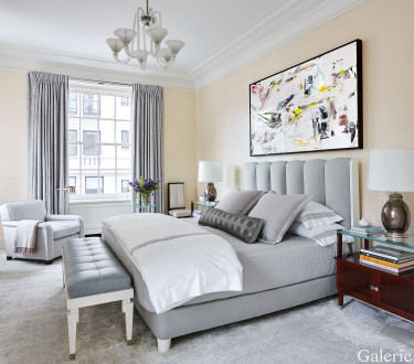 Elegant bedroom with a modern grey bed, abstract art on the wall, large window, chandelier, and cozy armchair.