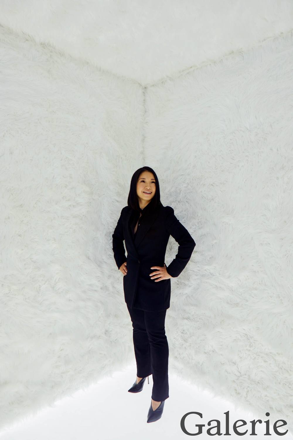 Person standing in white room with fur-textured walls, wearing a black suit and heels, facing forward and smiling.