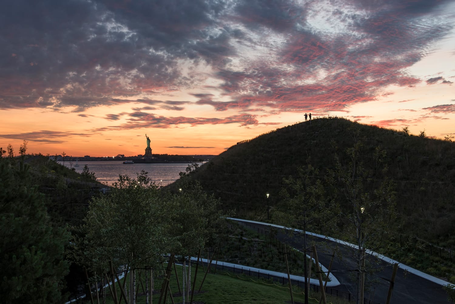 Sunset view of the Statue of Liberty from a hilltop with silhouetted figures and dramatic clouds in the background.