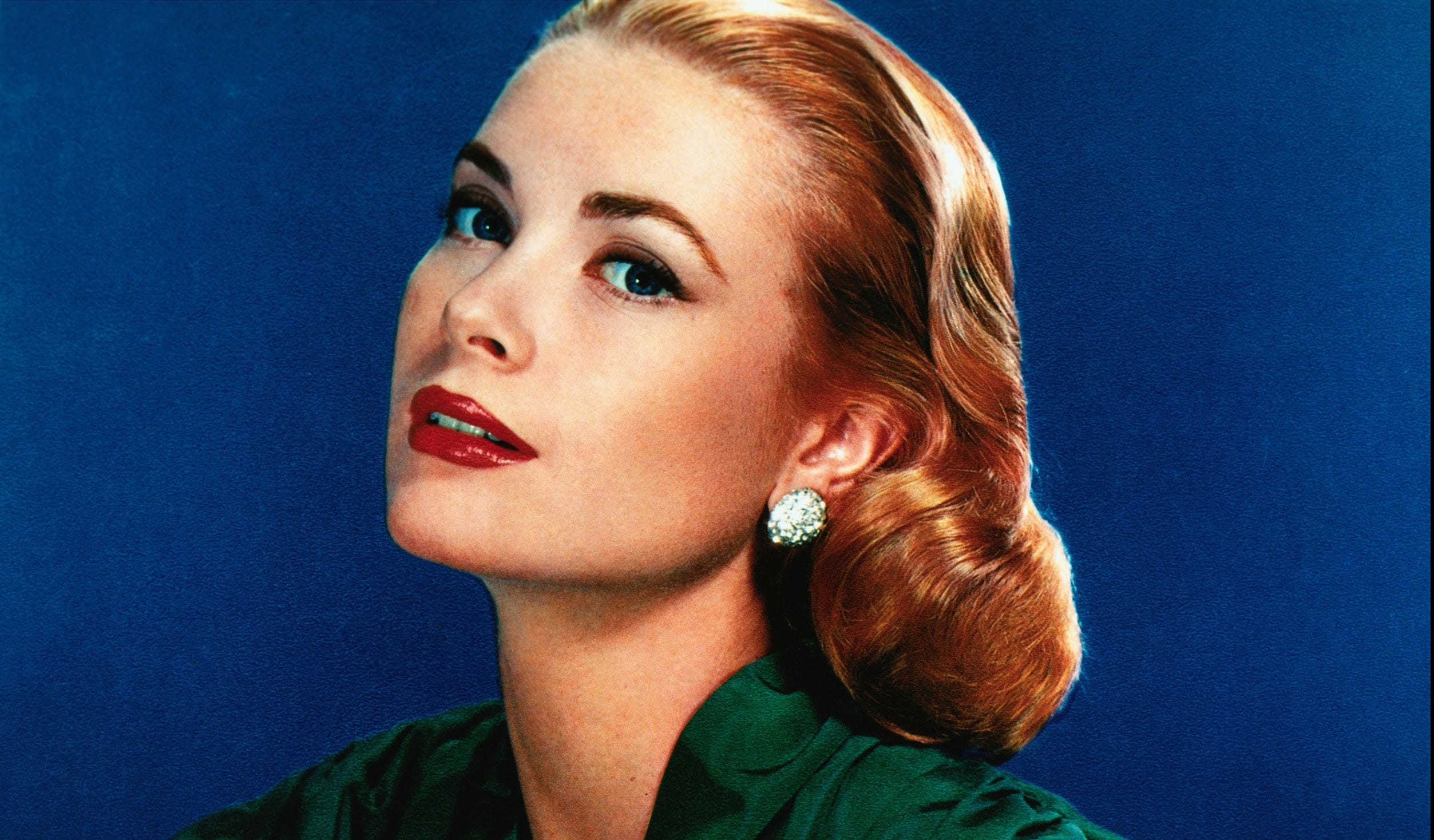 Woman with elegant hairstyle and earrings, posing against a blue background, wearing a dark green top and red lipstick