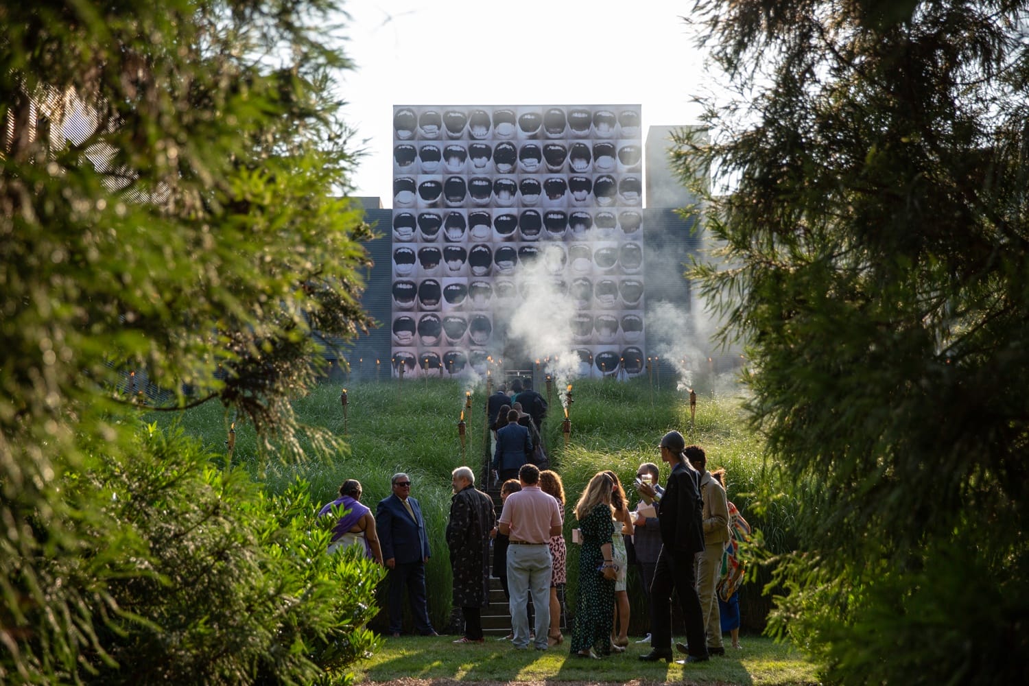 People walking towards a large, artistic installation outdoors surrounded by greenery and trees on a sunny day.
