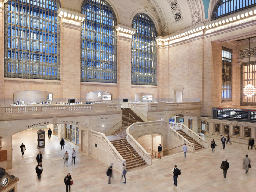 Grand Central Terminal interior with people walking, large windows, and elegant staircases in a historic architectural setting.