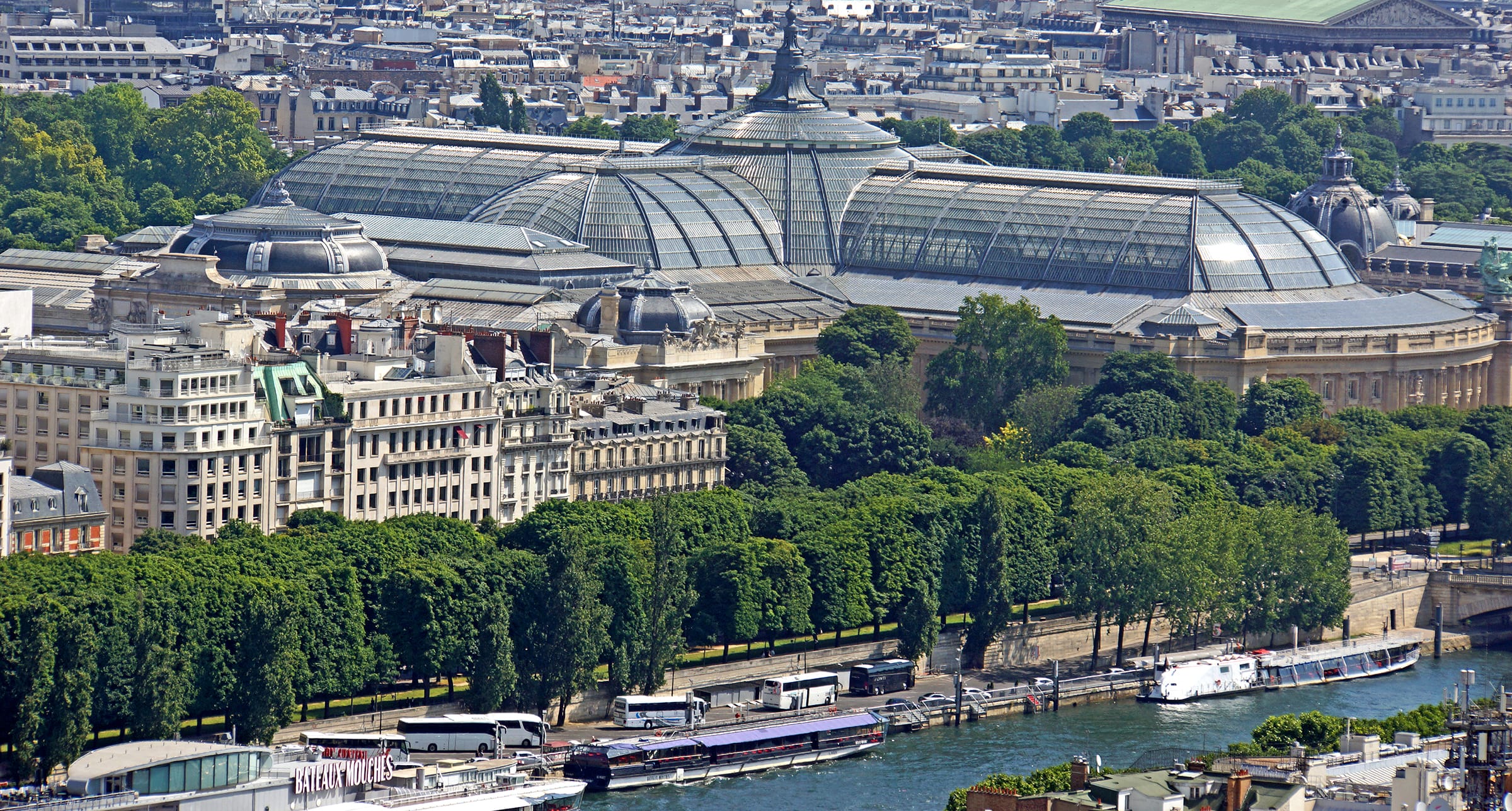 Aerial view of the Grand Palais in Paris with boats along the Seine River and surrounding trees.