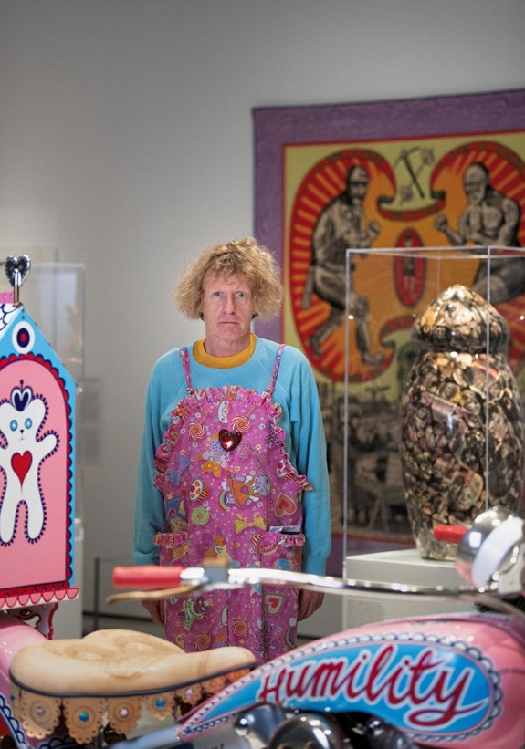 Person in colorful outfit standing in an art gallery with vibrant artworks and a decorated motorcycle in the foreground.
