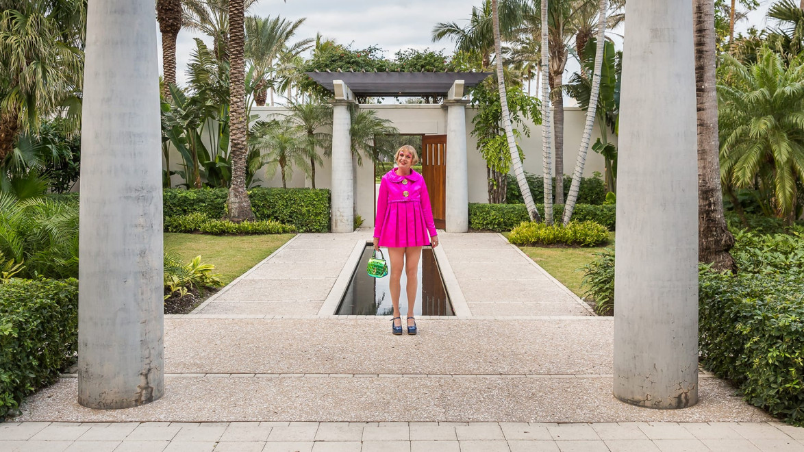 Person in a bright pink outfit stands between pillars in a tropical garden setting.