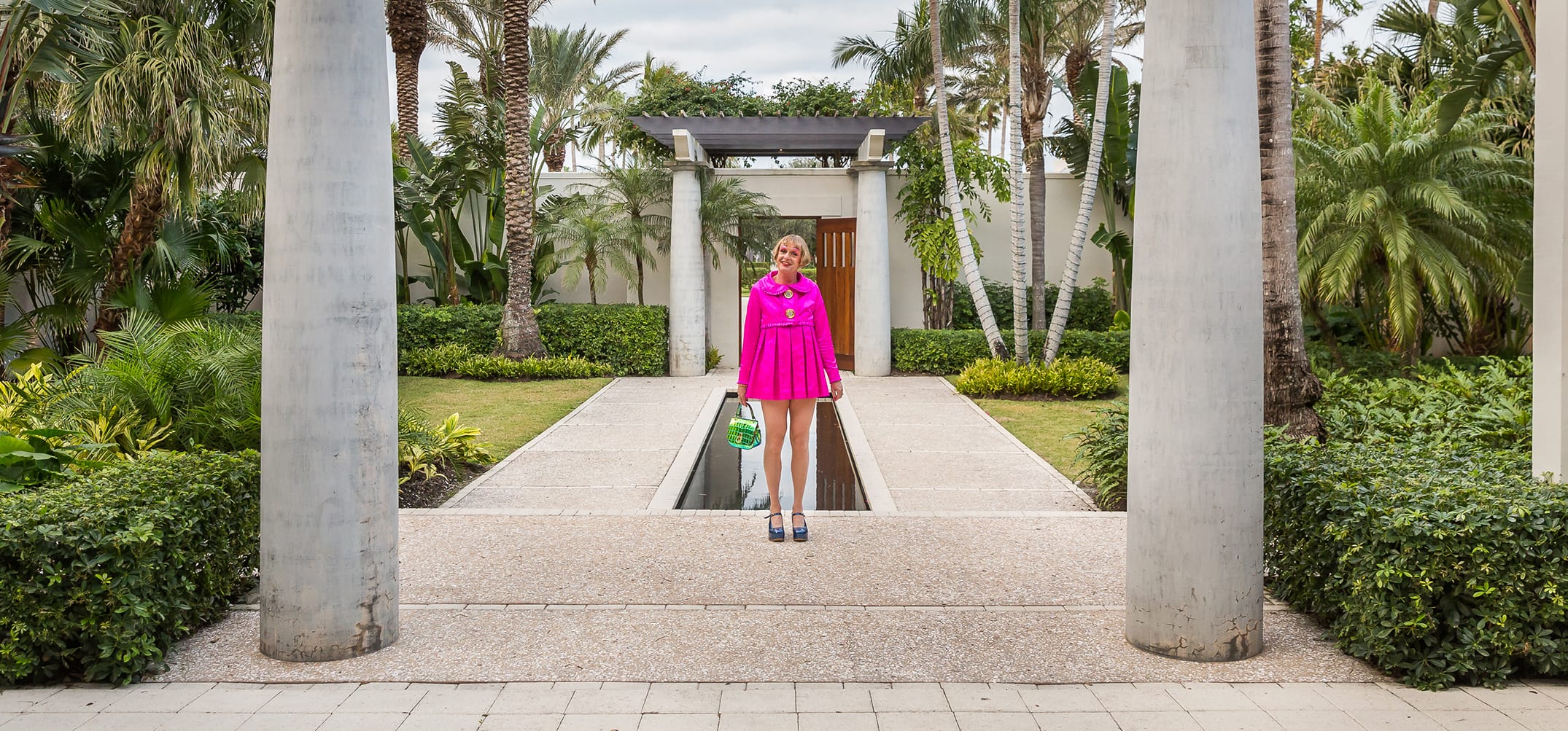 Person in a bright pink outfit stands between pillars in a tropical garden setting.