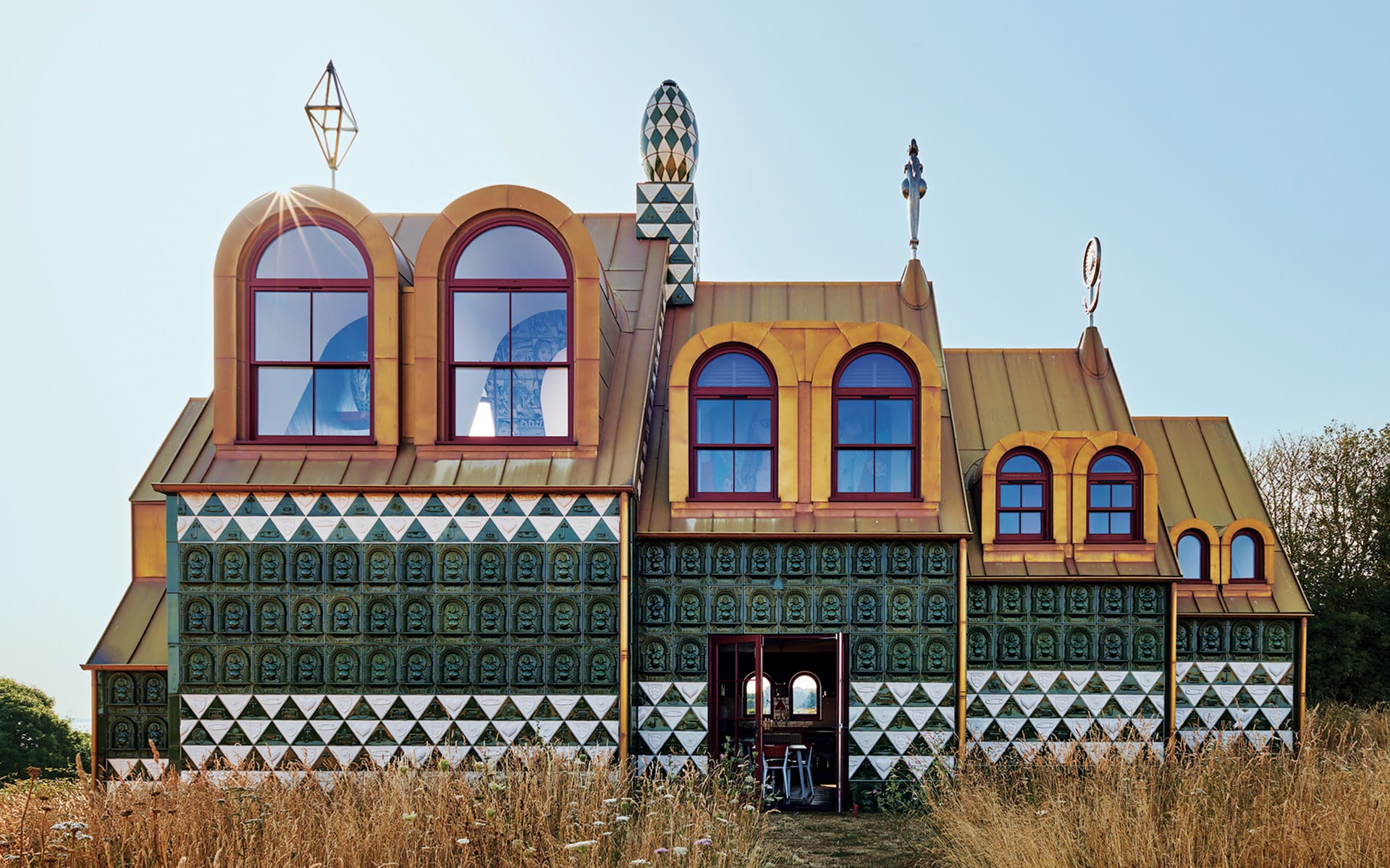 Unique, colorful house with geometric patterns, large arched windows, and artistic rooftop features surrounded by tall grass.