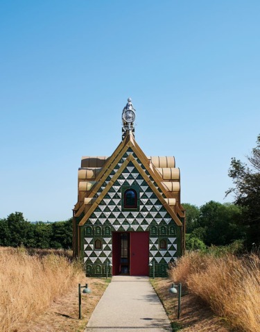 Colorful small chapel with geometric patterns and red door, set in a grassy landscape under a clear blue sky.