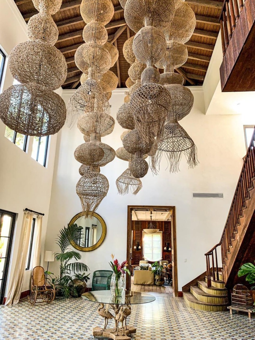 Spacious living room with high woven chandelier, wooden stairs, decorative plants, and patterned tile floor.