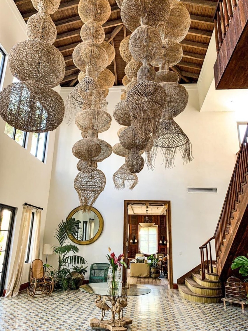Spacious living room with high woven chandelier, wooden stairs, decorative plants, and patterned tile floor.