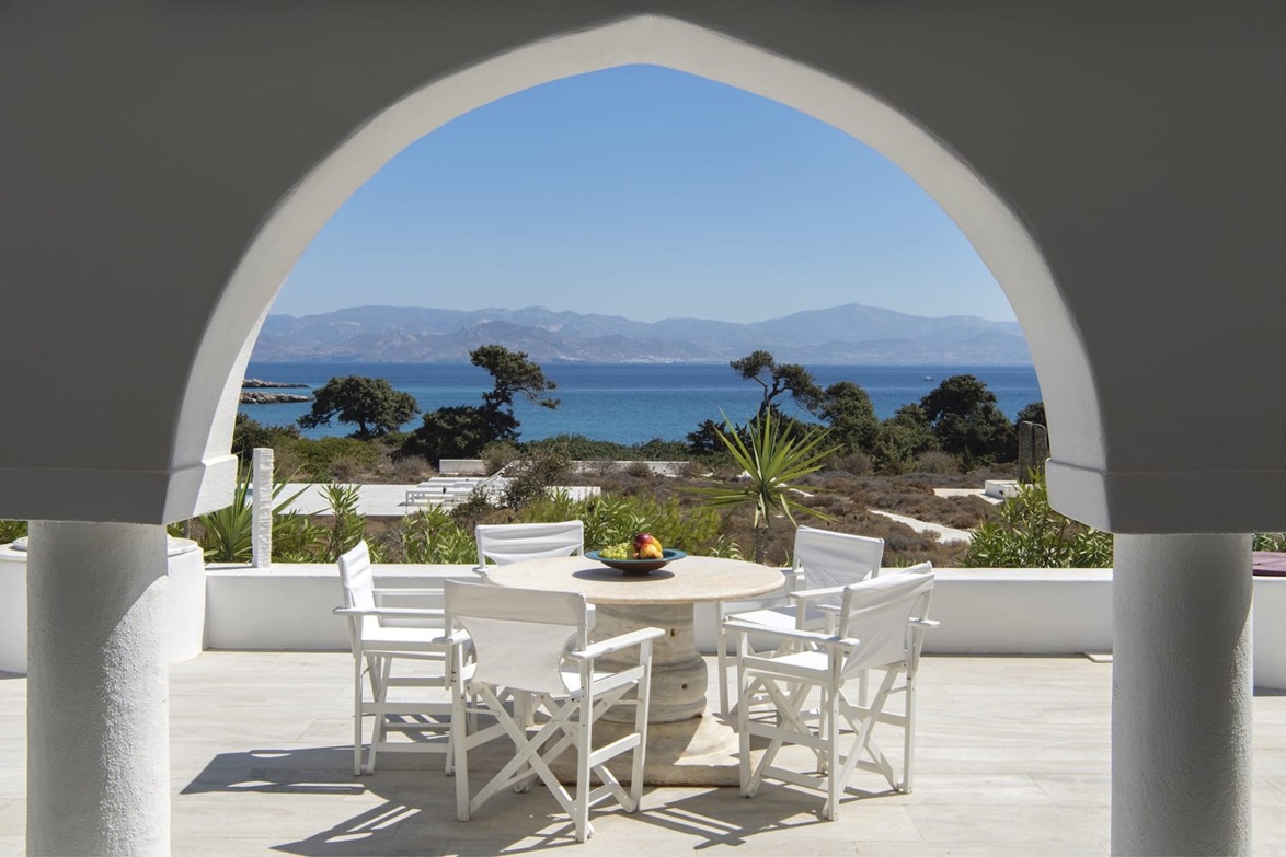 Arched veranda with white chairs and table overlooking sea and mountains in the distance on a sunny day.