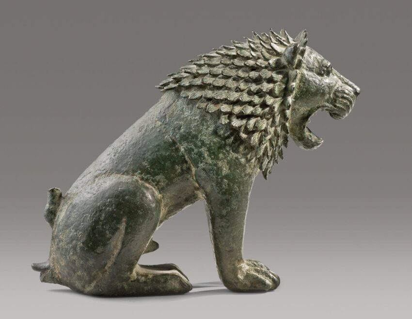 Ancient bronze lion sculpture with detailed mane, roaring posture against a plain background.