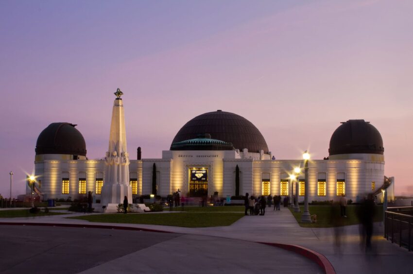 Griffith Observatory at sunset with lights illuminating the dome and tower, visitors walking nearby.