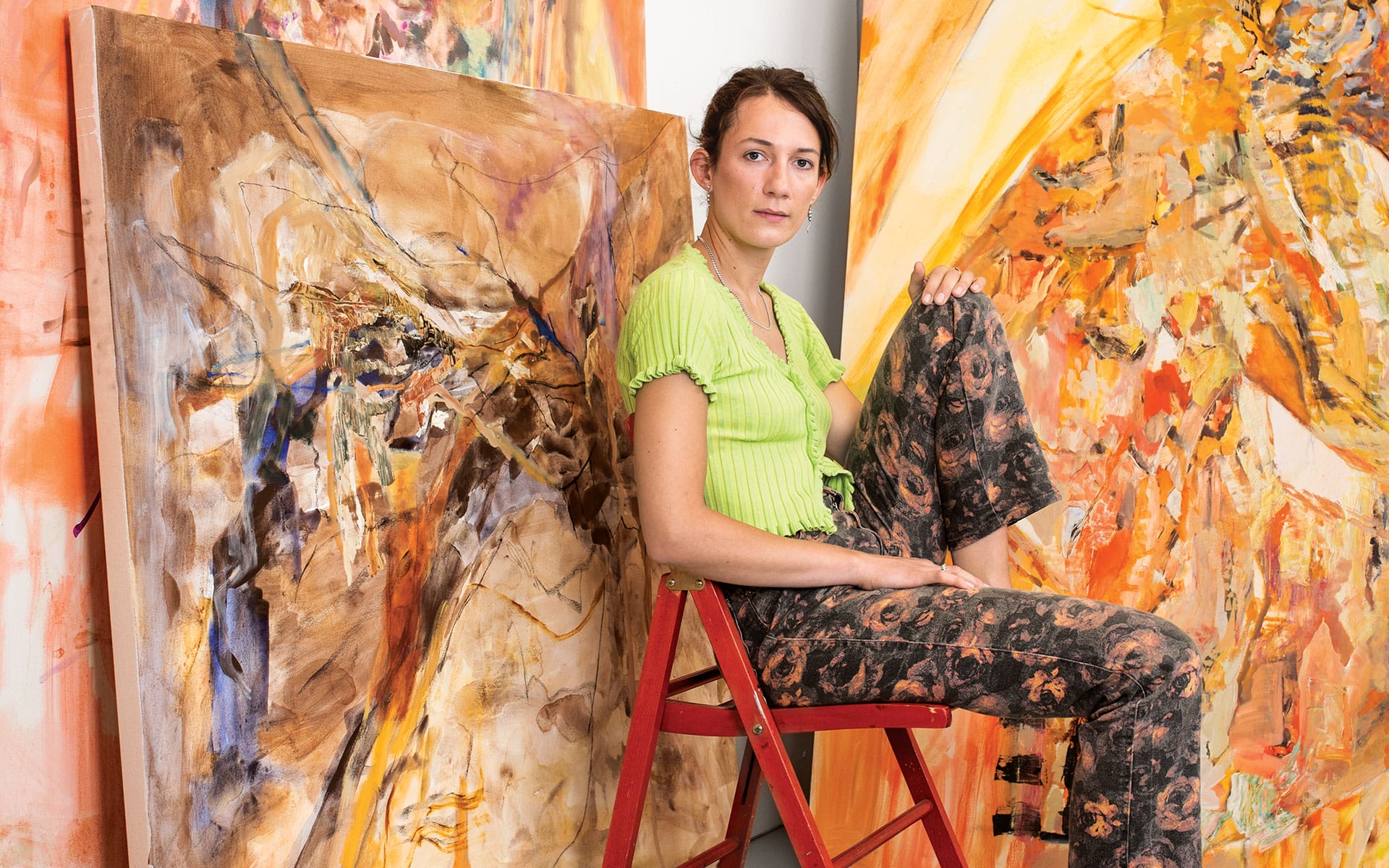 Artist sitting on a red ladder surrounded by large abstract paintings in a brightly lit studio.