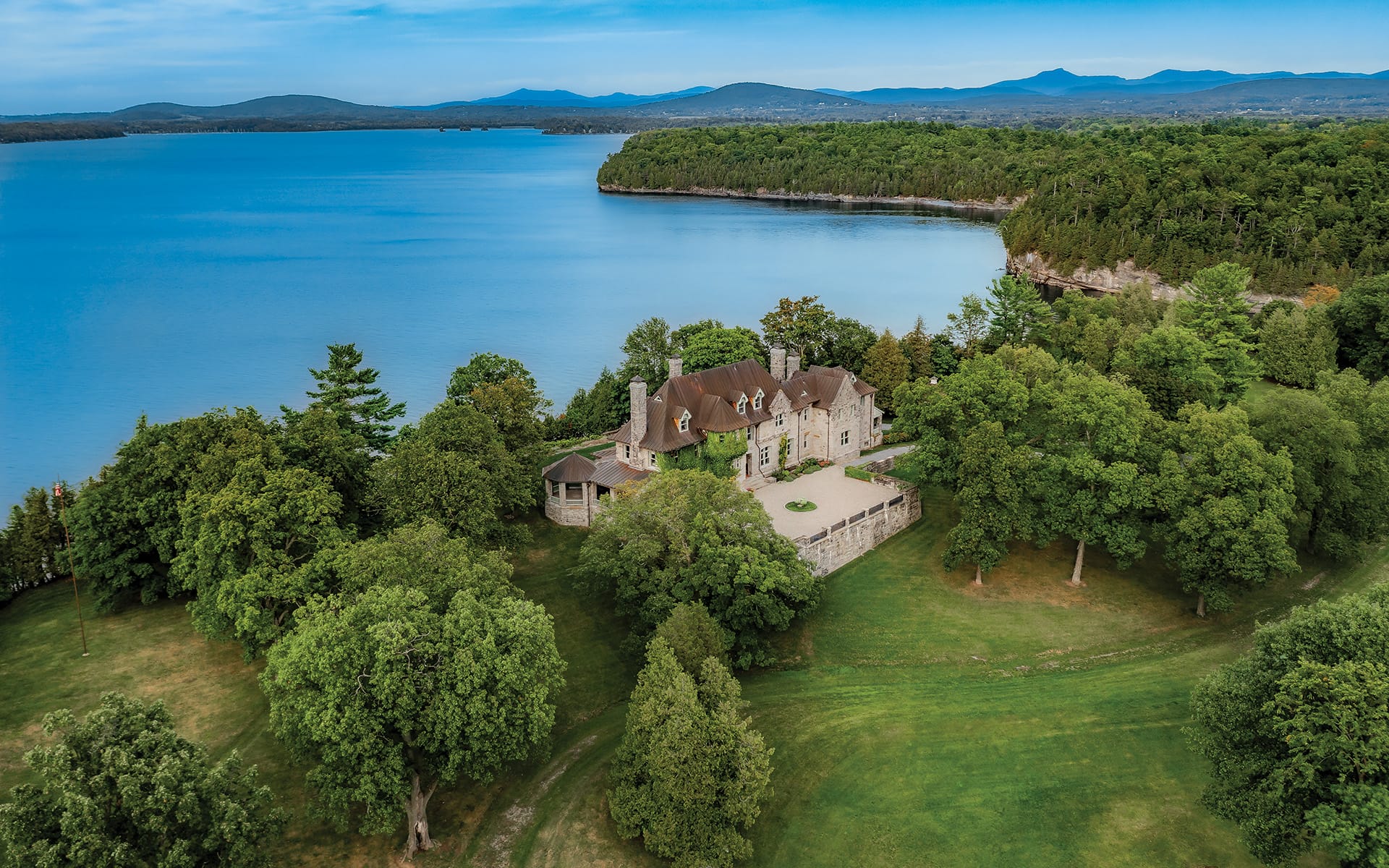 Aerial view of a large estate surrounded by lush greenery and overlooking a vast blue lake with distant mountains.