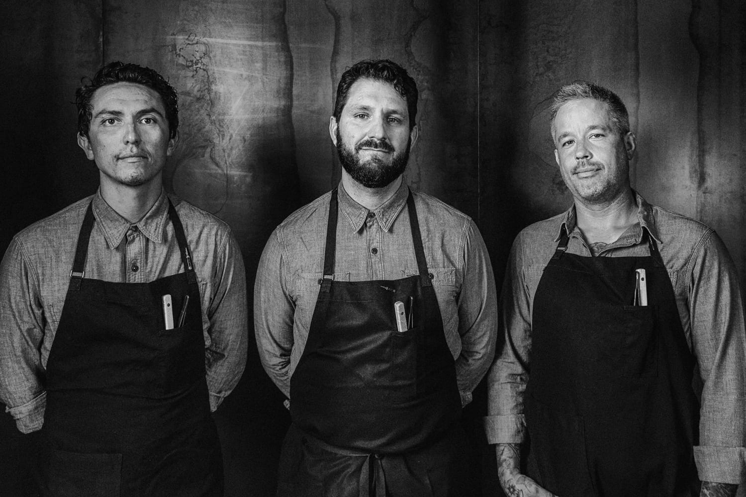Three chefs wearing aprons stand together, smiling, in a black and white portrait against a dark background.