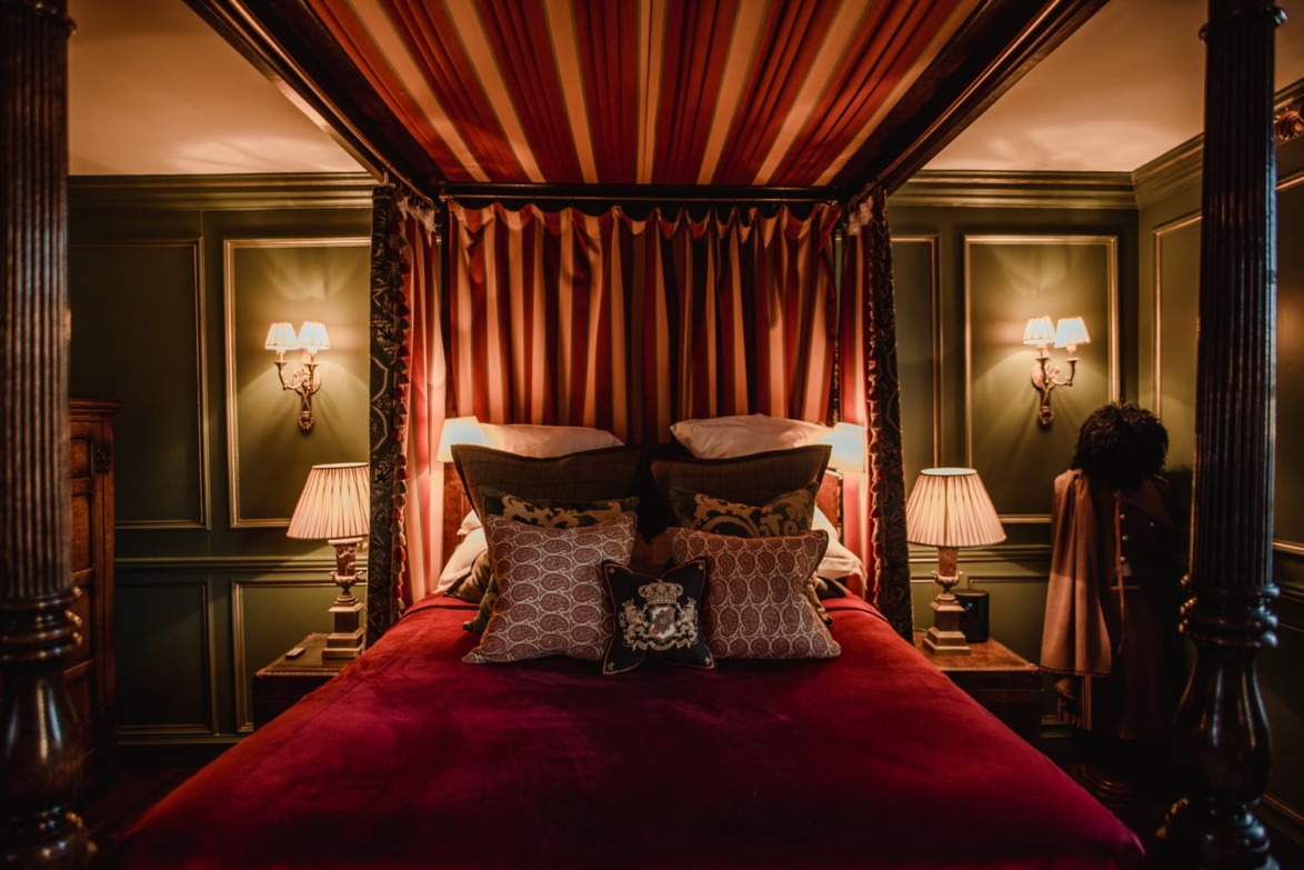 Luxurious bedroom with a four-poster bed, rich red bedding, ornate pillows, and soft lighting from wall lamps.