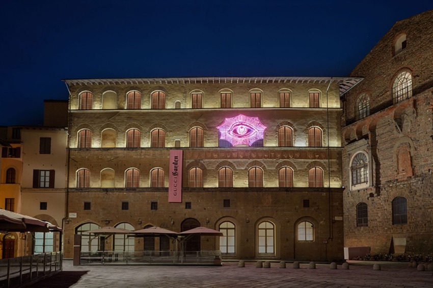 Illuminated historic building at night with a pink neon eye artwork on the facade in a city square.