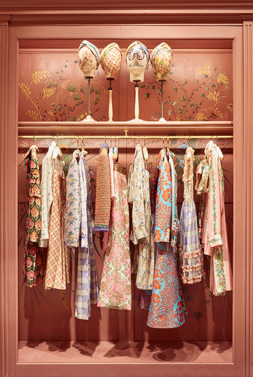 Vintage dresses on a rack with decorative hats above, set against a floral-themed backdrop in warm tones.
