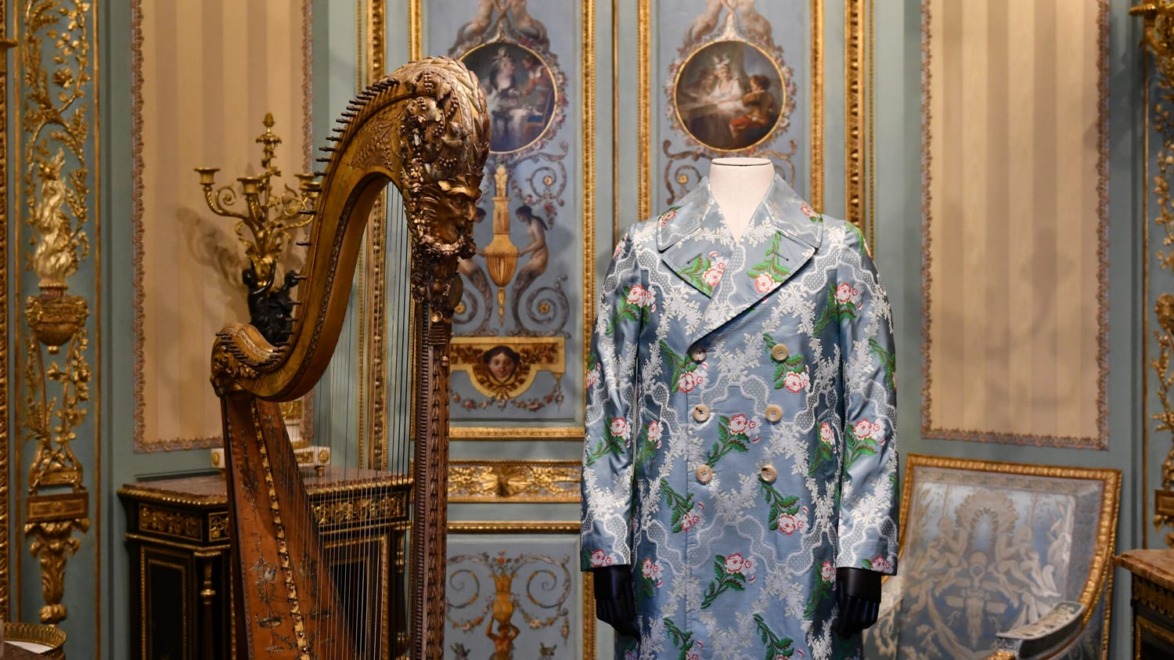 Elegant blue floral coat displayed in ornate room with harp and intricate decorations.