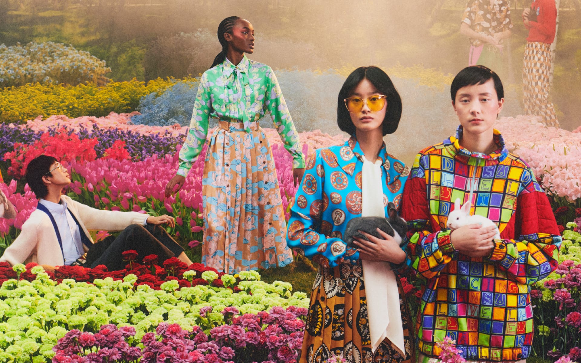 Group of models in vibrant clothing surrounded by colorful flowers, holding rabbits and showcasing diverse fashion styles.