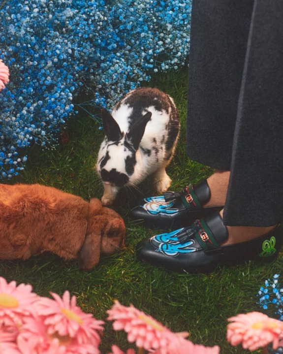 Person wearing shoes with bunny designs standing on grass with two rabbits surrounded by flowers.