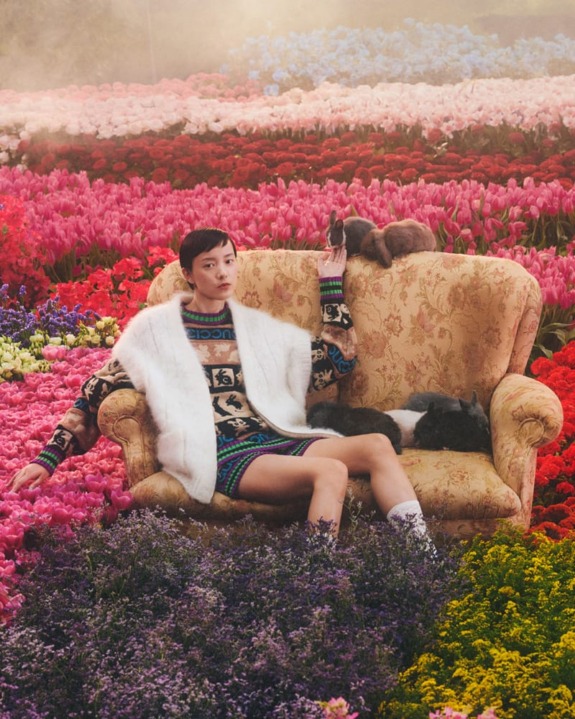 Person sitting on a floral couch among colorful flowers, wearing a patterned dress and white vest, with two rabbits nearby.