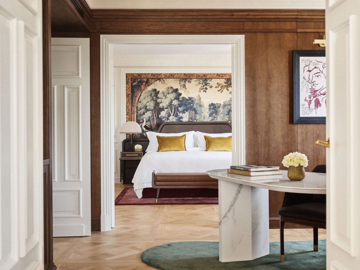 Elegant bedroom with a tapestry behind the bed, wooden walls, and a table with books and flowers in the foreground.