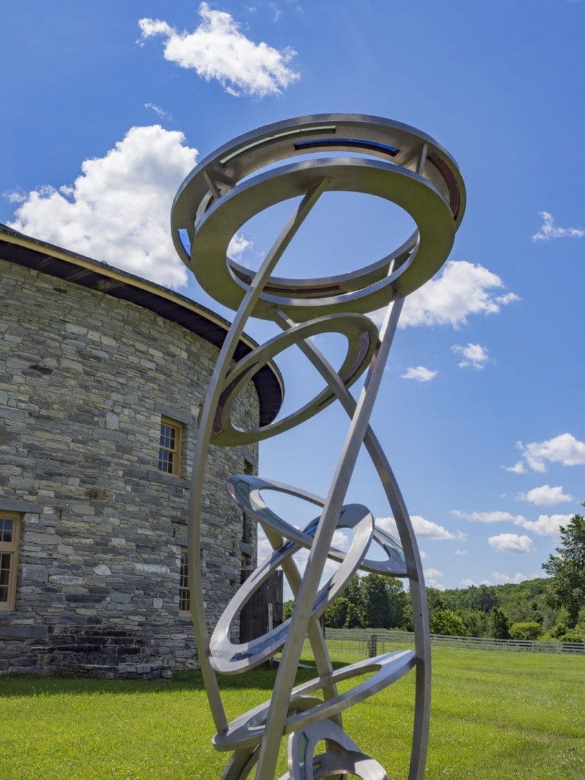 Modern metal sculpture with circular design near an old stone building, set against a backdrop of blue sky and clouds.