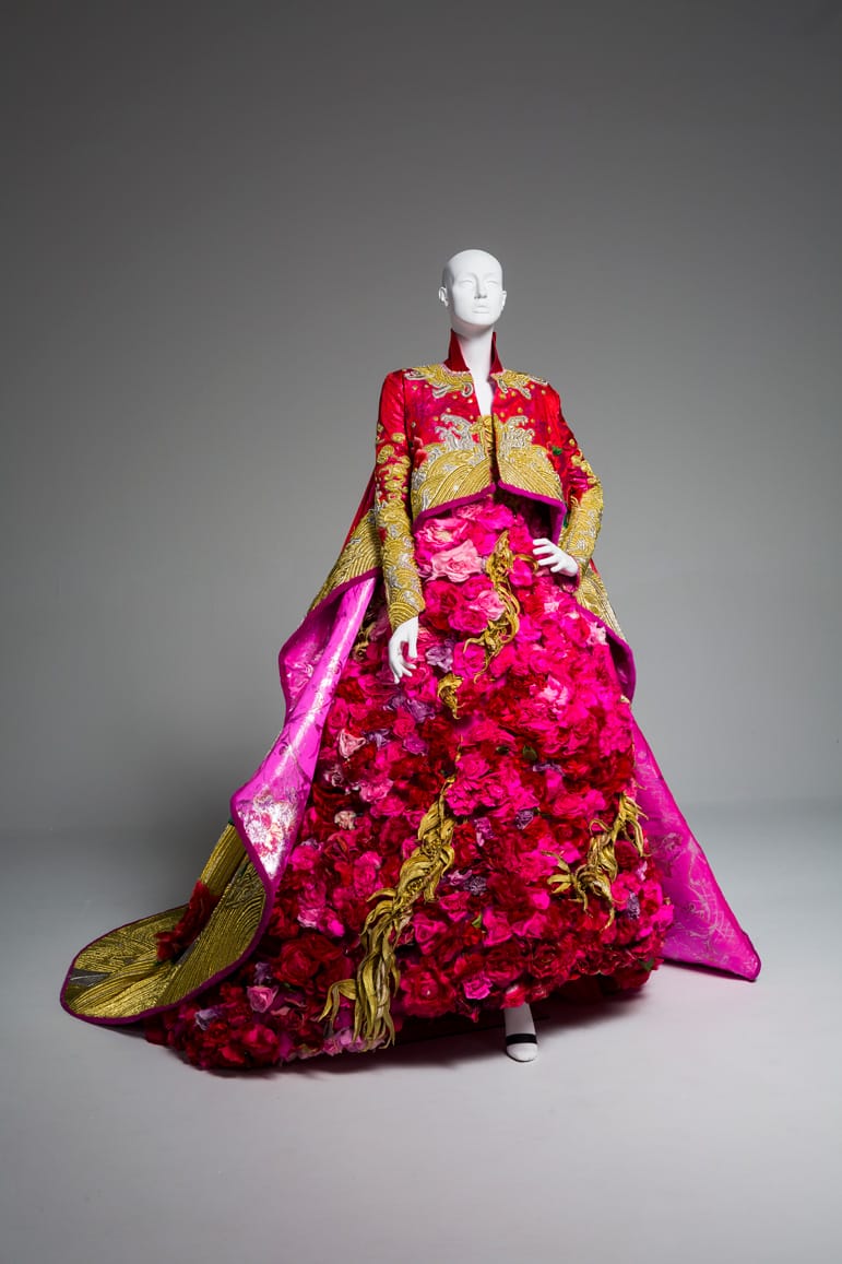 A mannequin wearing a vibrant pink and gold floral gown with intricate patterns and dramatic silhouette.