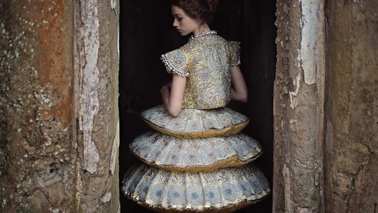 Woman in an ornate, layered dress standing in a rustic doorway.