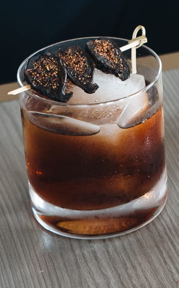 Cocktail in a glass with large ice cubes, topped with skewered dried figs on a wooden table.