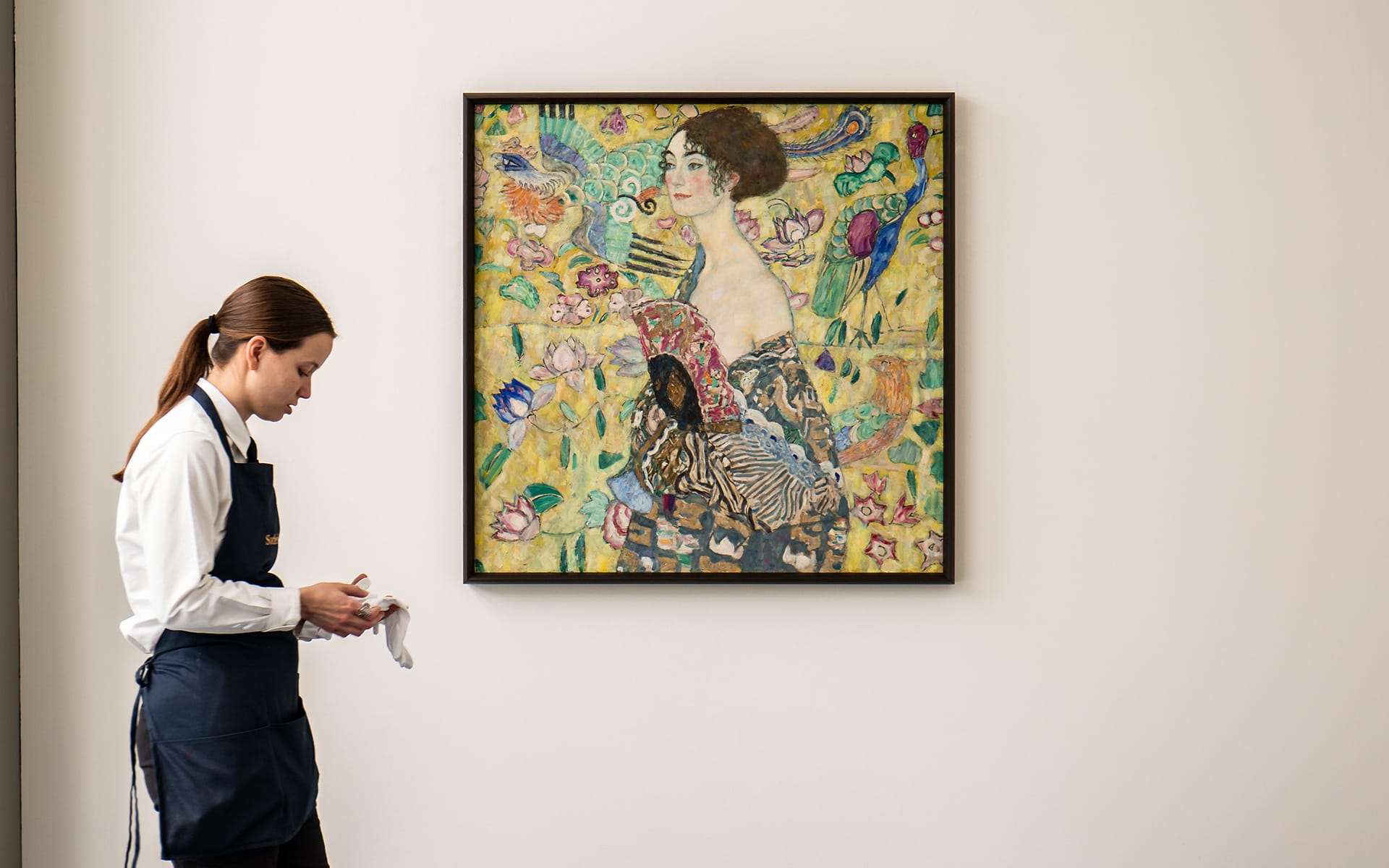 Museum staff member cleaning near a framed painting on a white wall.