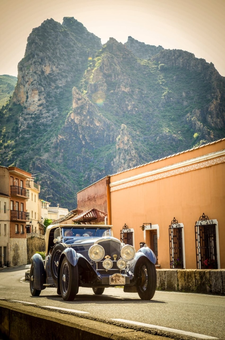 Vintage car driving through scenic village with mountain backdrop